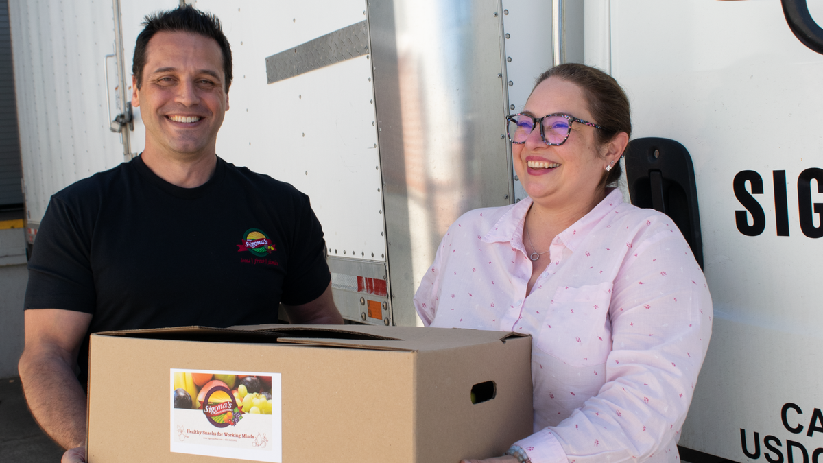 A man and a woman stand next to a white delivery truck holding a cardboard box with the text Sigona's Office Deliveries on it.